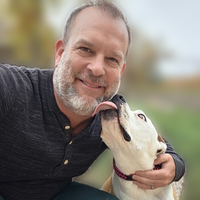 Arran Huckstep smiling outdoors while his dog licks his cheek, background softly blurred.